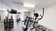 Located at 1722 N Walnut St in Bloomington, Indiana, this fitness room offers a well-organized exercise space designed to support a well-rounded workout routine. The room features durable rubber flooring throughout, providing a safe and practical surface for exercise. Bright flush-mounted ceiling fixtures ensure consistent, even lighting across the space.

The room is outfitted with a variety of equipment, including elliptical machines, a treadmill, and a multi-functional cable machine for strength training. A rack of dumbbells provides options for free weight exercises, while a water cooler and hand sanitizer station are conveniently positioned for user comfort and hygiene. A wall-mounted flat-screen television offers entertainment during workouts.

Full-length mirrors line one wall, visually expanding the room and allowing users to monitor their form. A large window with vertical blinds faces a green, wooded outdoor view, bringing in natural light and creating a pleasant exercise environment. A wall-mounted HVAC unit ensures climate control year-round. The clean, neutral white walls keep the atmosphere bright and focused. This fitness room delivers a comprehensive range of equipment and amenities within a compact, efficiently utilized footprint.