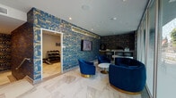 Located on Melrose Avenue in Los Angeles, this single-floor lobby and fitness center combines contemporary design with upscale finishes to create a welcoming and functional communal space. The lobby immediately impresses with its polished marble-look tile flooring, bold navy and gold abstract wallcoverings, and plush deep-blue velvet seating including a sweeping curved sofa and barrel chairs with gold-toned bases. A round marble-top coffee table anchors the lounge area, while floor-to-ceiling glass windows flood the space with natural light and offer open views of the tree-lined streetscape outside. A built-in workstation with a bar-height counter and rounded seating provides a practical workspace alongside the lounge. The elevator foyer features clean white paneled walls, a contrasting dark textured accent wall, and a sculptural circular chandelier that adds a refined focal point overhead. The on-site fitness center is thoughtfully equipped with TRUE brand cardio machines including treadmills, ellipticals, and stationary bikes, as well as a full dumbbell rack, a cable machine, an adjustable bench, and a stability ball. Motivational artwork and a wall-mounted television complete the gym environment. Throughout, recessed lighting and consistent marble-look flooring tie the spaces together cohesively.