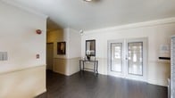 This single-floor space makes a strong first impression with its thoughtfully designed entryway. The foyer is anchored by a pair of elegant French doors that invite abundant natural light, framed by white-painted paneling and classic crown molding that lend a refined, traditional character to the space. Dark hardwood-style flooring runs throughout, providing a rich contrast against the crisp white walls and ceiling, creating a balanced and polished look. A decorative console table with a framed mirror serves as a welcoming focal point upon entry, adding a touch of curated style. The ceiling features clean lines with crown molding detail, contributing to a sense of height and openness. A corridor leads further into the interior, hinting at additional rooms beyond the entryway. The overall ambiance is bright, tidy, and well-maintained, with a neutral color palette that feels both inviting and versatile. The combination of classic architectural details, warm flooring, and generous natural light from the French doors gives the space a timeless, functional character suitable for a variety of uses.