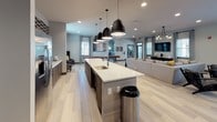 This single-story community amenity space is designed with a contemporary open-concept layout that seamlessly integrates multiple functional zones. Light hardwood-style flooring runs throughout, unifying the space and contributing to its bright, welcoming atmosphere. Natural light filters in through multiple windows dressed with floor-length curtains, complementing the recessed lighting overhead.

The kitchen area serves as a central hub, anchored by a generous white quartz island with an undermount sink and bar-height seating for several guests. Matte black industrial pendant lights hang above the island, adding a bold design contrast. Warm wood cabinetry and a stainless steel refrigerator line the back wall, alongside a textured mosaic tile accent wall framing a wall-mounted television — ideal for entertaining.

The open lounge area features plush neutral sectional sofas accented with yellow and patterned throw pillows, a wall-mounted TV, and an eclectic chandelier overhead. A game area along one side includes a foosball table and arcade cabinet, offering recreational options for residents. A separate dining nook with leather booth seating provides additional gathering space. Adjacent access leads to a fitness center, making this a well-rounded, multifunctional communal environment.