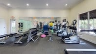 This single-floor fitness and recreation facility offers a comprehensive workout environment thoughtfully divided into distinct activity zones. The main gym area is generously sized and features a wide array of equipment, including treadmills, elliptical machines, a stationary bike, a multi-station strength training machine, free weights, benches, and balance training accessories. Large wall-mounted mirrors amplify the sense of space and allow users to monitor their form, while recessed lighting ensures a bright, well-lit environment. Several windows along one wall invite natural light and provide views of lush outdoor greenery, creating an energizing atmosphere. Carpeted flooring in the workout zones adds comfort underfoot, while the walkways feature durable tile. A mounted television is positioned for easy viewing during cardio sessions. Adjacent to the gym, a full-size racquetball court stands out as a premier feature of the facility. Enclosed by sleek glass walls on one side and smooth white walls on the others, the court boasts a polished hardwood floor with regulation court lines in red. The court's soaring ceiling height and overhead skylight panels ensure excellent visibility during play. A bulletin board and French doors near the gym entrance provide practical access and informational resources, completing this well-rounded athletic space.