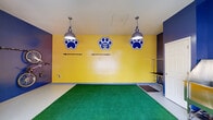 The Paw Spa is a purpose-built pet grooming facility designed with both functionality and a lively aesthetic in mind. The space is immediately recognizable by its bold color scheme — vivid yellow and deep navy blue walls adorned with oversized paw print logos — creating an energetic and welcoming environment for pets and their owners alike.

The main grooming area is the centerpiece of the space, featuring a stainless steel professional grooming tub equipped with steps for easy pet access, along with dedicated grooming tables mounted along the walls. Industrial-style pendant lights hang from the ceiling, providing ample illumination throughout the room. The floor is covered in bright green artificial turf, adding a playful, pet-friendly surface underfoot.

A wall-mounted bicycle rack serves as a practical storage solution, reflecting the space's efficient use of its footprint. Large floor-to-ceiling sliding glass windows and a classic paneled entry door flood the interior with natural light and offer clear views of the surrounding outdoor greenery and parking area, keeping the space feeling open and connected to the outside.

The facility also includes a utility area with rubber drainage mats near the grooming tub and wall-mounted access panels, ensuring the space meets the practical demands of professional pet care. Overall, the Paw Spa combines a fun, vibrant design with well-considered grooming amenities.