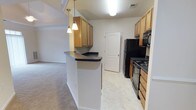 This single-floor residence offers a practical and inviting layout designed for comfortable everyday living. The kitchen serves as a central hub of the home, featuring warm honey-toned wood cabinetry that provides generous storage space. A peninsula with a dark countertop extends outward, creating a natural divide between the kitchen and the adjacent living area while offering additional prep and seating space. Pendant lighting hangs above the peninsula, adding a warm, focused glow to the workspace. The kitchen is equipped with a full suite of black appliances, including a refrigerator, range, and over-the-range microwave, all set against light tile flooring that is both durable and easy to maintain. A paneled interior door off the kitchen leads to additional space within the home. The adjoining living area transitions seamlessly from the kitchen, featuring plush carpeting that adds warmth and comfort underfoot. The overall palette of the space is neutral and clean, with white walls and standard ceiling heights that create a bright, airy atmosphere. The open flow between the kitchen and living area makes the space feel cohesive and well-connected, ideal for both everyday routines and casual entertaining.