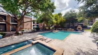 Located at 10864 Audelia Road in Dallas, Texas, this outdoor amenity area is the centerpiece of a well-maintained residential community. The space is anchored by a generously sized swimming pool with clear, turquoise water, framed by a stone-accented pool deck that provides ample room for lounging and recreation. Surrounding the pool, mature trees offer natural shade throughout the courtyard, creating a cool and inviting atmosphere even on warm Texas days. The multi-story brick residential buildings frame the space on multiple sides, featuring a classic design with blue accent paneling, balconies, and large windows that overlook the grounds. A dedicated patio area adjacent to the pool includes outdoor seating with umbrella-shaded tables and chairs, ideal for relaxing or socializing. A built-in BBQ grilling station is conveniently positioned nearby, making the space well-suited for outdoor gatherings. Colorful planters and neatly trimmed hedges add decorative touches throughout the landscaping, while stone-lined garden beds enhance the overall aesthetic. A gated entrance provides a defined boundary to the pool area, contributing to both safety and privacy. The overall ambiance is serene and resort-like, offering a welcoming outdoor retreat within an established Dallas neighborhood.