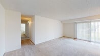 This single-floor unit offers a straightforward, functional layout with a clean, neutral aesthetic throughout. The living area is generously proportioned, featuring wall-to-wall carpet in a warm beige tone and a textured ceiling. Large vertical blinds cover a sliding glass door, allowing natural light to fill the space while providing access to the outdoors. An open sightline connects the living area to the adjacent dining space, which features hardwood-style flooring and a modest chandelier overhead.

The galley-style kitchen is efficiently arranged, featuring white cabinetry on both sides, a coil-burner electric range, a double-basin sink, and a full-size refrigerator. The hardwood-style flooring continues throughout, and under-cabinet lighting provides functional illumination. A hallway leads from the kitchen toward the rest of the unit.

The bedrooms are carpeted and finished with the same white walls found throughout the unit. One bedroom includes a closet with bifold doors, while another room features a window dressed with vertical blinds, offering natural light and ventilation through a ceiling vent. The bathroom includes a full tub-and-shower combination with chrome fixtures and white surround, alongside a toilet and hardwood-style flooring. Overall, the unit presents a clean, move-in-ready environment with consistent finishes throughout.