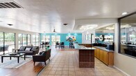 This single-floor community clubhouse is designed to foster social interaction and comfort through a well-planned, open layout. The main gathering area seamlessly blends a lounge and dining zone, featuring warm hardwood flooring that contrasts beautifully with classic tile accents. The lounge is furnished with plush sofas and accent seating, centered around a wall-mounted display, creating a relaxed and welcoming atmosphere.

The dining area is highlighted by a bold teal accent wall adorned with a vibrant multi-panel landscape artwork, setting a lively and energetic tone. High-top tables with granite surfaces and dark leather seating provide ample space for casual gatherings and social dining.

The kitchen is equipped with a full range, granite countertops, wooden cabinetry, and a commercial-style coffee station — ideal for hosting community events. A central island with a tiled base adds additional counter space and visual interest.

Large sliding glass doors and expansive windows flood the interior with natural light and open directly onto a generous outdoor deck, where umbrella-shaded seating overlooks a community pool surrounded by lush greenery. The transition between indoor and outdoor spaces is effortless, enhancing the overall sense of openness and connection to the outdoors.