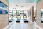 Located at 230 Bill Kennedy Way SE in Atlanta, GA, this well-appointed fitness center offers a thoughtfully designed exercise space across a single floor. The room features a contemporary aesthetic defined by light wood plank wall cladding, wood-toned flooring, and striking teal accent ceilings that add a bold pop of color against crisp white walls and structural columns.

Natural light floods the space through expansive floor-to-ceiling windows and glass doors that face an outdoor pool area, creating an open, energizing atmosphere. Linear LED lighting runs across the ceiling, ensuring the room remains bright and well-illuminated throughout.

The fitness floor is comprehensively equipped for a full range of workouts. A dedicated cardio zone features multiple treadmills positioned directly in front of the large windows, offering scenic outdoor views during use. Additional cardio options include elliptical machines and stationary cycling bikes with digital displays. A strength training area provides a wide selection of dumbbells, kettlebells, adjustable benches, and a cable machine. Exercise balls and a ballet barre round out the functional training offerings. Wall-mounted flat-screen televisions are positioned throughout the space for entertainment. Full-length mirrors along one wall visually expand the room, while a wooden bench near the entrance provides a convenient seating area.