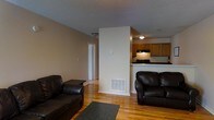 This single-floor residence offers a practical and comfortable layout with an open-concept design that connects the living area to the kitchen. The living room is a welcoming space, furnished with dark leather seating arranged around a low-profile area rug, and finished with warm hardwood flooring that extends throughout the unit. Neutral beige walls and a textured ceiling contribute to a calm, understated ambiance. A central partial-wall divider separates the living space from the kitchen while maintaining an open sightline, adding a sense of architectural definition without closing off the floor plan. The kitchen features warm-toned wood cabinetry, dark countertops, and a freestanding range with an overhead ventilation hood. A raised counter along the kitchen's edge provides a casual bar-style seating opportunity and further connects the cooking and living areas. A floor vent provides in-unit climate control. A hallway visible from the living area leads to additional rooms, suggesting a functional multi-room layout. The hardwood floors, consistent wall tones, and efficient use of space make this unit a straightforward and livable single-floor home.