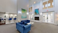 Located at 601 Rosery Rd NE in Largo, Florida, this single-floor commercial space offers a generously sized, open-concept interior designed with a clean, contemporary aesthetic. The most striking architectural feature is the dramatically high ceiling, which lends an expansive, airy quality to the entire space and is complemented by large upper-level windows that flood the interior with natural light.

The main area is defined by a neutral color palette of crisp whites and soft grays, with plush gray carpeting underfoot providing warmth and comfort throughout. Tall, structural columns add architectural interest while naturally dividing the open floor plan into functional zones. A built-in bar counter with bold blue bar stools serves as a focal point near the entry, creating an inviting, social atmosphere. A set of French doors opens to the exterior, enhancing connectivity and allowing additional natural light to pour in.

The seating areas are thoughtfully arranged with glass-top round tables paired with sleek white chairs, while a pair of deep blue tufted sofas anchors a lounge zone alongside a white fireplace. Decorative ceramic urns, curated artwork, and accent lighting further elevate the refined ambiance. The space also features a dedicated service or reception counter area, making it well-suited for professional or community use.