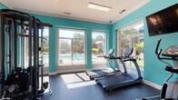 The fitness center at 2500 Grove Club Lane in Cary, NC is a well-appointed, single-floor facility designed to support a comprehensive workout experience. The space is immediately distinguished by its bold teal walls accented with yellow striping, white crown molding, and rubber-floored workout zones that lend the room an energetic yet clean aesthetic.

Large, tall windows line multiple walls throughout the facility, flooding the interior with natural light and offering direct views of the outdoor pool area and surrounding landscaped grounds. The openness created by the windows makes the space feel airy and inviting.

The fitness center is organized into distinct functional zones. A cardio area features treadmills, elliptical trainers, and a stationary bike, with wall-mounted flat-screen televisions positioned for easy viewing during workouts. A dedicated free weights section includes a full dumbbell rack with a range of weights, exercise balls, medicine balls, and a pull-up bar mounted to the wall. The strength training area houses a multi-functional cable machine and additional weight equipment. A stretch and flexibility zone offers step platforms and stability balls alongside tall windows overlooking the pool.

Mirrored walls in key sections enhance the sense of space and provide visual feedback during exercise. Ceiling fans and recessed lighting ensure comfort throughout. The overall ambiance is vibrant, functional, and well-lit — a comprehensive community fitness amenity.
