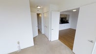 This single-floor residence offers a clean and functional layout with a bright, neutral aesthetic throughout. The interior is characterized by crisp white walls and a combination of soft carpeting in the living and hallway areas, transitioning to warm hardwood-style flooring in the open-concept living and kitchen space.

The kitchen is positioned at the heart of the home, visible through an open passthrough that connects it seamlessly to the main living area. It features dark countertops, white cabinetry, and stainless steel appliances including a refrigerator and microwave, giving it a modern and practical feel. The open layout encourages a natural flow between cooking and living spaces.

The hallway area provides access to additional rooms and includes a set of bi-fold closet doors, offering generous storage solutions. Flush-mount ceiling lighting keeps the spaces well-lit throughout. The overall ambiance is calm and understated, with a move-in ready quality that makes the space highly versatile. The consistent use of white tones and neutral flooring creates a cohesive look that is both timeless and adaptable to a variety of interior styles.