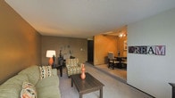 This single-floor space is designed with an open-concept layout that connects the living area seamlessly to the dining space, creating a functional and cohesive flow. The living room is carpeted and features warm, earthy-toned walls complemented by a textured ceiling. A large window with vertical blinds allows natural light to fill the room, enhancing its comfortable atmosphere. The space accommodates a plush sofa and patterned accent chair arranged around a dark wood coffee table, creating a welcoming seating area.

Transitioning from the living room, a distinctly painted accent wall in a rich orange hue marks the entrance to the dining area. This space features a ceiling fan with a light fixture overhead and is furnished with a dining table and chairs, making it suitable for everyday meals. Decorative wall art adds a personalized touch to the dining space.

Throughout the unit, neutral carpet flooring unifies the rooms, while the combination of warm and neutral tones on the walls contributes to a cozy, lived-in ambiance. The layout is practical and well-proportioned, offering clearly defined areas for living and dining within a single connected floor plan.