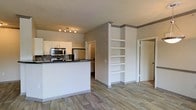 This single-floor space showcases an open-concept layout that seamlessly connects the kitchen and living areas. The kitchen is equipped with stainless steel appliances, including a microwave, stove, and refrigerator, complemented by white cabinetry and a dark granite-style countertop that extends into a breakfast bar peninsula. A mosaic tile backsplash adds a decorative touch behind the cooking area, while a track lighting fixture overhead ensures ample illumination.

The living area features light hardwood-style flooring that runs consistently throughout the space, contributing to a cohesive and airy feel. A standout architectural detail is the built-in white shelving unit nestled into a partial wall divider, offering both functional storage and a display element. The neutral warm-grey wall tones provide a versatile backdrop throughout.

Two doorways are visible — one leading to an additional room or hallway toward the back, and another framed door to the right, suggesting further living or storage spaces. The overall ambiance is clean, modern, and practical, with a neutral palette that allows for flexible interior styling.