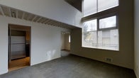 This two-story space is defined by its dramatic double-height ceiling in the main living area, which creates a strong sense of openness and volume. A mezzanine level is visible overhead, bordered by a metal railing, adding architectural depth and a sense of vertical scale. Large windows along one wall flood the space with natural light while offering views of surrounding greenery, enhancing the connection to the outdoors. The windows are fitted with horizontal blinds, providing privacy and light control. The main level features wall-to-wall carpeting in a neutral gray tone, contributing to a subdued, functional aesthetic. Exposed structural elements, including beams on the upper portion of the wall, add an industrial character to the interior. An adjacent room is accessible through an open doorway and features track lighting on the ceiling, suggesting its use as a functional secondary space. The overall design balances utilitarian elements with architectural interest, making it a versatile space suited for a variety of uses. Neutral wall tones throughout maintain a clean, understated atmosphere.