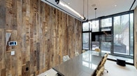 This thoughtfully designed single-floor space masterfully combines rustic charm with contemporary industrial aesthetics. The centerpiece of the room is a striking full-height reclaimed wood accent wall, showcasing a rich mosaic of weathered planks in varying tones and textures that add warmth and character to the space. Opposite the wood wall, expansive floor-to-ceiling black-framed windows flood the interior with abundant natural light while offering views of lush greenery outside, creating a seamless connection between the indoors and the natural environment. The room is anchored by a sleek, large-format conference table with a glossy surface, surrounded by elegantly patterned upholstered chairs that introduce a decorative contrast to the room's industrial elements. Overhead, a pair of pendant light fixtures suspended by black chains provide focused task lighting, complementing the exposed track lighting mounted to the ceiling. A wall-mounted flat-screen display is integrated into the windowed wall, enhancing the space's functionality as a professional meeting environment. The combination of raw materials, refined furnishings, and generous daylighting creates an atmosphere that is both productive and visually compelling.