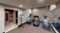This single-floor fitness facility offers a practical and well-organized space for cardiovascular exercise and wellness. The room features neutral-toned walls and carpeted flooring with dark wood baseboards, creating a clean and functional environment. Recessed ceiling lighting provides steady, even illumination throughout.

The main exercise area is outfitted with a selection of cardio equipment, including a treadmill, elliptical trainer, air resistance bikes, and a stair climber, each positioned on protective floor mats. A large floor-standing fan is available to aid ventilation during workouts. One wall is lined with a full-length mirror, visually expanding the space and allowing users to monitor their form during exercise.

A standout feature of this facility is the adjoining sauna room, accessible through a wooden door, offering a dedicated space for post-workout relaxation and recovery. An additional door leads to a separate room, providing further utility within the facility. Wall-mounted décor, including a bicycle-themed art piece, adds a subtle fitness-inspired touch to the space. Safety is addressed with clearly posted emergency contact signage. A wall-mounted HVAC unit ensures climate control. Overall, this fitness space is thoughtfully arranged to accommodate a range of exercise needs in a comfortable, accessible setting.