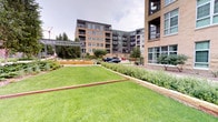 This thoughtfully designed outdoor courtyard garden sits nestled within a contemporary multi-story brick and glass residential development, offering a well-maintained green retreat in an urban setting. The centerpiece of the space is a broad, immaculately kept lawn bordered by raised planting beds filled with a diverse mix of shrubs, ornamental grasses, perennials, and ground cover plants, creating a rich tapestry of greenery and texture throughout the landscape.

Defined garden edges, accented with corten steel-style borders, give the space a clean and structured aesthetic while maintaining a natural, organic feel. A metal pergola structure anchors the midground of the garden, providing a focal architectural element and a shaded gathering point within the landscape. Mature trees frame the perimeter of the space, adding vertical scale and a sense of established, natural character.

The surrounding multi-story buildings, constructed with warm brick facades and featuring private balconies with large windows, enclose the courtyard on multiple sides, creating a sense of privacy and seclusion despite the urban context. The overall ambiance is serene and inviting, offering residents and visitors a tranquil outdoor environment that balances structured landscape design with natural plantings and open green space.