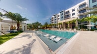 This striking outdoor amenity area is defined by its generously sized resort-style swimming pool, which serves as the centerpiece of the space. The pool features sleek, modern in-water lounge chairs that invite relaxation just beneath the surface, creating a signature architectural detail that evokes a luxury resort atmosphere. The surrounding deck is finished in light-toned pavers, providing a clean and polished aesthetic that complements the shimmering turquoise water.

Lining the pool deck is an array of cushioned lounge chairs and white shade umbrellas, offering ample space for sunbathing and outdoor leisure. Towering palm trees are thoughtfully interspersed throughout the area, providing a lush tropical backdrop and natural shade. The landscaping adds a vibrant, verdant layer to the overall ambiance, enhancing the sense of a private oasis.

The pool area is framed by a modern multi-story building with a contemporary façade featuring clean lines, private balconies, and a neutral color palette accented with teal tones. A covered pavilion or cabana structure is also visible, providing additional shaded gathering space. The entire setting is bathed in abundant natural sunlight under an open sky, reinforcing the bright, open, and resort-inspired character of this outdoor amenity space.