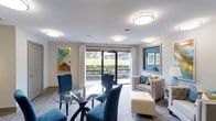 This WiFi area is a thoughtfully designed communal space that blends comfort and functionality in a bright, inviting atmosphere. The single-floor layout is illuminated by multiple flush-mount ceiling lights that cast an even, warm glow throughout the room. A large sliding glass door serves as a focal point, framing a verdant outdoor view of manicured greenery and allowing natural light to flood the interior. Sheer patterned curtains flank the door, adding a soft, elegant touch.

The seating arrangement is both practical and stylish. A round glass-top dining table surrounded by plush, deep-blue upholstered chairs creates an ideal spot for working or connecting with others. Complementing this is a cozy lounge area featuring a cream-toned settee and a tufted ottoman, accented with colorful throw pillows that echo the space's vibrant color palette.

Vivid artwork adorns the walls, with an abstract, multi-colored canvas on one side and a framed blue-toned piece on the other, lending a contemporary artistic character to the space. The neutral walls and carpeted floors provide a calm backdrop that allows the bold furnishings and décor to stand out. Overall, this WiFi area offers a polished, comfortable environment well-suited for both productivity and relaxation.