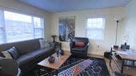 This single-floor space offers a comfortable and stylishly designed interior that balances modern aesthetics with a welcoming ambiance. The living room serves as the centerpiece of the space, bathed in natural light streaming through multiple large windows fitted with horizontal blinds. Light-toned walls create a clean, airy backdrop that complements the room's bold design choices. The flooring features warm hardwood tones that anchor the space with a sense of warmth and continuity. A plush gray sofa and a sleek black leather armchair, accented with a deep red bolster pillow, provide ample seating arranged around a wooden coffee table. A striking black-and-white large-format cityscape photograph — depicting a dramatic aerial view of a metropolitan skyline — serves as the room's focal point, adding an urban, cosmopolitan energy to the interior. The space is further enlivened by a bold geometric patterned area rug in black, red, and blue tones, which adds texture and visual interest underfoot. Decorative accessories, including sculptural objects on the coffee table and a floral arrangement, contribute to the curated, intentional feel of the space. Overall, this single-floor space presents a thoughtfully designed environment that feels both stylish and functional.