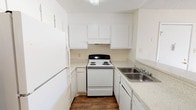 This single-floor space includes a well-organized kitchen designed for efficient use of available room. The kitchen features a clean, all-white aesthetic, with white upper and lower cabinetry providing ample storage throughout. A white electric coil-top range with a black control panel serves as the focal point, positioned beneath a built-in overhead exhaust hood. A full-size white refrigerator stands to the left, complementing the cohesive color scheme. To the right, a double-basin stainless steel sink is set into a speckled laminate countertop that extends along the perimeter, offering a practical workspace. A dishwasher is also integrated beneath the counter near the sink. The flooring throughout the kitchen consists of dark wood-style planks, providing a warm contrast to the bright white cabinetry and walls. Recessed lighting above keeps the space well-lit. The layout follows a U-shaped configuration, maximizing the use of the available footprint while maintaining clear pathways. The overall design is straightforward and functional, with a neutral palette that creates a clean and approachable environment.
