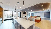 This thoughtfully designed single-floor amenity space offers a generous, open-plan layout that seamlessly connects a fully equipped kitchen, a combined kitchen and living room, and a welcoming entrance area. The kitchen is anchored by a substantial island with a crisp white quartz countertop, dark cabinetry, and a full row of bar seating — all illuminated by a series of sleek pendant lights. Open shelving with integrated accent lighting and a full suite of stainless steel appliances add both function and style. The living area is centered around a linear gas fireplace set against a textured tile surround, with a wall-mounted television above. Multiple seating configurations — including a curved banquette nook with pendant lighting and various lounge groupings — create versatile spaces for gathering or relaxing. Warm wood flooring, a sophisticated blue-grey ceiling detail with cove lighting, and floor-to-ceiling black-framed windows and doors fill the interior with natural light. The entrance features a built-in niche with floating shelves and backlit accent lighting. Direct access leads to two outdoor terraces: a courtyard-facing lounge area and an open-air deck with panoramic neighborhood views, both furnished for outdoor living.
