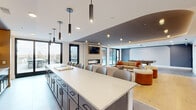 This thoughtfully designed single-floor amenity space offers a generous, open-plan layout that seamlessly connects a fully equipped kitchen, a combined kitchen and living room, and a welcoming entrance area. The kitchen is anchored by a substantial island with a crisp white quartz countertop, dark cabinetry, and a full row of bar seating — all illuminated by a series of sleek pendant lights. Open shelving with integrated accent lighting and a full suite of stainless steel appliances add both function and style. The living area is centered around a linear gas fireplace set against a textured tile surround, with a wall-mounted television above. Multiple seating configurations — including a curved banquette nook with pendant lighting and various lounge groupings — create versatile spaces for gathering or relaxing. Warm wood flooring, a sophisticated blue-grey ceiling detail with cove lighting, and floor-to-ceiling black-framed windows and doors fill the interior with natural light. The entrance features a built-in niche with floating shelves and backlit accent lighting. Direct access leads to two outdoor terraces: a courtyard-facing lounge area and an open-air deck with panoramic neighborhood views, both furnished for outdoor living.
