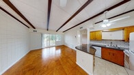 This single-floor unit offers a thoughtfully designed layout that blends character and functionality throughout. The main living area immediately draws the eye upward to its striking exposed dark wood beam ceiling with a tile panel finish, which lends a warm, architectural charm to the space. Gleaming parquet hardwood floors extend throughout the living and bedroom areas, adding richness and continuity to the interior. Ceiling fans are positioned in both the living area and kitchen, ensuring comfortable airflow throughout.

The kitchen is open and efficiently arranged, featuring warm honey-toned wood cabinetry, a dark granite countertop peninsula that separates the kitchen from the living space, a tile backsplash, and white appliances including a range, microwave, refrigerator, and dishwasher. Light tile flooring grounds the kitchen zone and transitions cleanly from the hardwood in the adjacent living area.

The living room is generously proportioned, with access to an outdoor balcony through sliding glass doors that invite natural light and views of surrounding greenery. A dedicated bedroom offers parquet flooring, a window with outdoor views, and a distinctive tiled accent wall. The full bathroom is clean and functional, appointed with a vanity, mirror cabinet, tub-shower combination, and tile flooring.