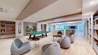This generously sized amenity lounge is designed to deliver a sophisticated, multi-functional social experience across a single open floor. The space is accessed through a striking curved entryway with a circular recessed ceiling detail, setting an elegant tone before entering the main area. Light-toned hardwood flooring flows throughout, complementing the clean, contemporary aesthetic defined by neutral tones and warm wood accents.

At the heart of the space sits a full-size pool table, anchoring a casual games and lounge zone furnished with distinctive rounded pod-style chairs and sleek black side tables. Built-in open shelving units line the walls, offering display and storage space, while potted tropical plants add organic warmth. A dedicated media lounge features a large wall-mounted flat-screen television set against a warm cognac-toned accent wall, flanked by floor-to-ceiling built-in bookshelves and comfortable white upholstered seating arranged on a patterned area rug.

Expansive floor-to-ceiling windows flood the lounge with natural light and frame views of an outdoor terrace. A kitchenette area is appointed with veined white marble-style countertops and backsplash, stainless steel appliances, and minimalist cabinetry. A dedicated workspace with ergonomic task chairs rounds out the functional offerings. Recessed lighting throughout ensures a well-lit, inviting ambiance at any hour.