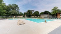 This outdoor amenity space centers around a generously sized rectangular swimming pool with clean, clear blue water and classic metal entry railings. The surrounding concrete deck offers ample room for relaxation, lined with multiple lounge chairs that invite residents to unwind in the open air. A striped umbrella provides a shaded retreat on sunny days, adding a pop of color to the clean, neutral tones of the patio surface. The pool area is fully enclosed by a perimeter fence, ensuring a safe and defined recreational zone. Mature, leafy trees frame the space on all sides, creating a natural canopy that offers partial shade and a serene, park-like atmosphere. The lush green lawn beyond the fence reinforces the open, airy feel of the environment. Multi-story residential buildings are visible in the background, situating this pool as a shared community amenity within a larger residential setting. The overall ambiance is bright, welcoming, and well-maintained, making it an ideal outdoor gathering and leisure destination during warmer months.