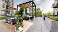 Located at 6550 York Ave S in Edina, Minnesota, this thoughtfully designed outdoor amenity space serves as a central gathering hub for residents. The terrace spans a generous footprint and is framed by a multi-story building with a contemporary facade combining brick, corrugated metal cladding, and private balconies that overlook the courtyard below.

The space is organized into several distinct zones, each designed for relaxation and socializing. Multiple steel-framed pergolas with warm wood-slat canopies are anchored throughout, strung with café lights that create an inviting ambiance. Beneath the pergolas, curated seating arrangements feature sectional sofas, lounge chairs, and accent tables suited for both casual conversation and unwinding outdoors.

Lush artificial turf panels introduce a vibrant green contrast against the neutral tones of the large-format tile pavement that extends across the terrace. Raised concrete planters filled with ornamental grasses and flowering plants add natural texture and greenery throughout. A cornhole board and additional outdoor game areas encourage recreational activity.

Perimeter railings open to views of the surrounding Edina streetscape and mature trees, connecting the space visually to its neighborhood context. The overall design balances urban sophistication with comfort, creating an outdoor retreat that is both functional and aesthetically refined.
