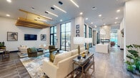 Located at 320 S Walker St in Cary, NC, this single-floor amenity space presents a sophisticated, contemporary design characterized by soaring ceilings, an open layout, and carefully curated interiors. The welcoming reception area features a teal-accented desk beneath a sculptural globe chandelier, setting an upscale tone from the entry.

The main lounge areas flow seamlessly into one another, offering multiple seating arrangements with neutral-toned sofas, accent chairs, and layered area rugs. Linear pendant lighting and recessed fixtures illuminate the space, complementing the abundant natural light streaming through floor-to-ceiling black-framed windows and glass doors. These expansive windows provide direct views and access to an outdoor patio area.

A striking stacked-stone fireplace serves as a focal point in one lounge zone, flanked by a billiards table and additional gathering spaces. Curated artwork and decorative accents add visual interest throughout. A kitchen-style bar area with teal cabinetry and marble countertops provides functional service space. The varied flooring—combining large-format tile and wood-look planks—helps define distinct zones within the open-plan layout, creating a cohesive yet dynamic communal environment.