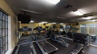 This fitness center is a fully equipped, single-floor exercise facility designed to support a comprehensive workout routine. The room features a dark rubber flooring throughout, providing a durable and slip-resistant surface ideal for a variety of exercises. The textured ceiling is fitted with fluorescent lighting that ensures the space is well-lit, while large multi-pane windows along one wall allow natural light to filter in and offer views of an outdoor pool area, creating a refreshing workout environment. Full-length mirrors line one side of the room, enhancing the sense of space and allowing users to monitor their form during exercise. The cardio section is well-appointed with multiple treadmills and elliptical machines, offering ample options for aerobic training. On the strength training side, the facility includes a multi-function cable machine, a dedicated weight bench, a dumbbell rack with a range of free weights, and a collection of medicine balls in various sizes. A padded adjustable seat is also available for targeted exercises. Motivational fitness posters are displayed on the walls, adding an energetic atmosphere. The space is accessed through a standard door and benefits from a climate control vent in the ceiling, ensuring comfort throughout the year.