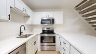 This two-story condo spans 1,200 square feet, with 790 sq ft on the main floor and 410 sq ft upstairs. The open main level features a combined living room and kitchen with light hardwood flooring throughout. The kitchen includes white shaker cabinets, a white peninsula, and stainless steel appliances. A staircase with a metal railing leads to the upper level, where the 280 sq ft primary bedroom and additional bedrooms are located, each featuring ceiling fans and windows. The bathrooms include tub-shower combinations. A dedicated laundry room houses a full-size washer and dryer. A balcony provides views of the courtyard and community pool below.