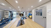 This fitness facility offers a thoughtfully designed workout space spread across a single level, delivering a clean and functional environment for a comprehensive exercise experience. The room is finished with light hardwood flooring that lends warmth and brightness throughout, complemented by recessed ceiling lighting that ensures the space is evenly and efficiently illuminated.

Large windows along one wall invite natural light and provide views of the surrounding greenery, creating an open and energizing atmosphere. A full wall-length mirror amplifies the sense of space while allowing users to monitor their form during workouts. One section of the room features a bold teal accent wall, adding a vibrant visual contrast to the otherwise neutral palette of grey and white tones.

The facility is equipped with a variety of cardio machines, including stationary bikes and a treadmill, as well as a multi-function cable strength machine and an assortment of free weights. A dedicated rack holds multiple stability and exercise balls in varying sizes. Motivational fitness posters and a workout chart are displayed on the walls, enhancing the purposeful atmosphere. A wall-mounted flat-screen television provides entertainment during workouts, and a separate door offers convenient access to adjacent areas.