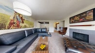 Located on W University Street in Wichita, Kansas, this single-floor residence offers a well-designed layout that balances comfort and functionality throughout. The expansive living room serves as the centerpiece of the home, featuring plush carpeting, a tiled fireplace with a wood mantel, and a wall-mounted television. Large sliding glass doors open to a private balcony furnished with bistro seating, bringing in natural light and outdoor views of the surrounding community. The adjacent dining area is distinguished by dark exposed ceiling beams and a stylish pendant light fixture, creating a warm and defined space for entertaining. The kitchen connects seamlessly to the dining area via a peninsula counter with a built-in sink, complemented by dark cabinetry, a range, and a refrigerator, all set over hardwood-style flooring. The primary bedroom is generously sized with carpeted floors and ample room for furnishings, while an additional bedroom offers playful versatility with space for a loft bed setup. The bathrooms are finished with rich wood-toned vanities, stone-look tile flooring, and a full tub-shower combination. Throughout the residence, textured ceilings and neutral wall tones provide a clean, cohesive aesthetic.