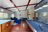 This inviting community lounge in Marlborough, MA, offers a well-appointed gathering space defined by striking architectural details and a warm, social atmosphere. The interior is anchored by soaring vaulted ceilings adorned with rich, dark wood exposed beams that lend a sense of grandeur and character throughout. Rich hardwood flooring runs the length of the space, complementing the neutral gray walls and bold blue accent features.

A floor-to-ceiling stacked stone fireplace serves as the room's dramatic focal point, rising prominently and framed by a dark wood mantel. Multiple seating arrangements — including plush sofas, armchairs in deep blue tones, and casual accent chairs — are thoughtfully arranged to encourage both relaxed lounging and social interaction. A full-size pool table with pendant lighting overhead adds an entertainment element to the space.

Natural light pours in through large windows fitted with sheer curtains, offering pleasant views of the surrounding greenery. A compact kitchenette area is visible toward the rear of the space, adding convenience for gatherings. Glass doors open to an outdoor balcony, further connecting the interior to the natural surroundings. The lounge is a versatile, well-designed communal retreat suited for both quiet relaxation and lively recreation.