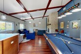 This inviting community lounge in Marlborough, MA, offers a well-appointed gathering space defined by striking architectural details and a warm, social atmosphere. The interior is anchored by soaring vaulted ceilings adorned with rich, dark wood exposed beams that lend a sense of grandeur and character throughout. Rich hardwood flooring runs the length of the space, complementing the neutral gray walls and bold blue accent features.

A floor-to-ceiling stacked stone fireplace serves as the room's dramatic focal point, rising prominently and framed by a dark wood mantel. Multiple seating arrangements — including plush sofas, armchairs in deep blue tones, and casual accent chairs — are thoughtfully arranged to encourage both relaxed lounging and social interaction. A full-size pool table with pendant lighting overhead adds an entertainment element to the space.

Natural light pours in through large windows fitted with sheer curtains, offering pleasant views of the surrounding greenery. A compact kitchenette area is visible toward the rear of the space, adding convenience for gatherings. Glass doors open to an outdoor balcony, further connecting the interior to the natural surroundings. The lounge is a versatile, well-designed communal retreat suited for both quiet relaxation and lively recreation.