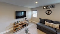 This single-floor space is thoughtfully designed with a cohesive modern aesthetic, featuring light hardwood-style flooring and neutral warm-gray walls that create a clean, inviting atmosphere throughout. Recessed lighting provides consistent illumination across all areas, complementing the natural light that filters in through windows along the exterior-facing walls.

The main lounge area is generously proportioned, offering ample room for relaxed seating. A comfortable sectional sofa is paired with mid-century modern accent chairs arranged alongside a small side table, while framed black-and-white photography adorns the walls, lending a curated, artistic character to the space. Potted plants, including a large fiddle-leaf fig, add organic warmth and texture.

A secondary seating nook features a wall-mounted flat-screen television above a rustic wood media console, creating a casual entertainment zone with a welcoming feel highlighted by decorative signage and a classic analog clock.

The dedicated workstation area accommodates multiple users at a long wooden counter-height table equipped with desktop computers and a printer, making it well-suited for productivity. Framed artwork continues the gallery-style theme here as well.

Overall, the space balances functional utility with stylish, contemporary design details throughout.