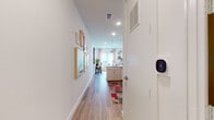 This single-floor residence is designed with a clean, modern aesthetic characterized by crisp white walls, recessed lighting, and light-toned hardwood-style flooring that flows throughout the main living areas. A welcoming entryway hallway sets the tone upon entry, lined with gallery-framed artwork and equipped with a smart thermostat for climate control convenience.

The kitchen is efficiently designed, featuring white shaker-style cabinetry extending to the ceiling, white quartz countertops, and a marble-look tile backsplash. Stainless steel appliances — including a range, over-the-range microwave, dishwasher, and refrigerator — complete the space. Pendant lighting adds warmth above the kitchen island area.

The living room is bright and airy, benefiting from multiple large windows dressed with blush pink curtains and horizontal blinds that allow ample natural light. A ceiling fan provides added comfort, while the open layout easily accommodates both a living and dining area.

The bedroom offers plush carpeting, a spacious walk-in closet, and a distinctive black-hardware sliding barn door. Neutral walls provide a calm backdrop, and the room connects seamlessly to the closet and additional storage. Overall, the unit presents a cohesive, contemporary living environment.