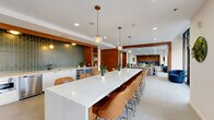 Located at 160 Morgan St in Jersey City, NJ, this expansive single-floor amenity space offers a generously sized, open-plan layout designed for both socializing and relaxation. The entrance greets visitors with large-format porcelain tile flooring, bold black-framed glass doors, and curated artwork, setting a sophisticated tone throughout.

The kitchen and dining area serve as the centerpiece of the space, anchored by a striking elongated white quartz island with seating for multiple guests. The kitchen wall features a full suite of stainless steel appliances, a matte black sink, a built-in wine refrigerator, and a distinctive sage-green ceramic tile backsplash framed by warm wood cabinetry. Pendant lighting with brass and globe accents adds warmth overhead.

The living room offers a versatile lounge environment with plush seating, mid-century-inspired leather chairs, and layered area rugs over carpeted flooring. A warm wood-paneled accent wall defines a more intimate meeting or dining nook, equipped with black ergonomic chairs. Floor-to-ceiling windows along one wall flood the space with natural light and offer views of a landscaped outdoor terrace. Black-framed glass partitions create semi-private zones while preserving the open, airy feel of the overall layout.
