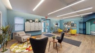 This single-floor residential lobby makes an immediate impression with its bold teal and light blue accent walls set against crisp white ceilings outfitted with sleek linear LED lighting. Wood-look plank flooring runs throughout, lending warmth and continuity to the space. The lobby is organized into several distinct functional zones. Near the entrance, a bank of individual resident mailboxes lines the wall alongside a secure glass entry vestibule, providing convenient mail access and controlled building entry. Adjacent to the entrance, a well-appointed lounge area features an eclectic mix of mid-century modern seating — including vibrant yellow accent chairs, a blue sofa, and charcoal wingback chairs — arranged atop colorful floral area rugs. Gold-toned open shelving units display curated décor, framed artwork, and books, adding character to the space. A secondary seating nook near a striking arched iron gate offers a more intimate gathering area, with a round floral rug anchoring a pair of grey chairs and a compact sofa. The ornate iron gate serves as an architectural focal point, referencing classic building heritage. Teal-framed elevator doors and doorways reinforce the cohesive color palette throughout. Potted plants placed throughout the lobby add organic freshness. Overall, the space balances functional building amenities with a lively, design-forward aesthetic.
