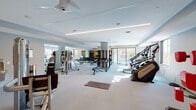 This contemporary fitness center is designed to support a full range of workout routines within a thoughtfully organized, single-level layout. The space is finished with neutral gray walls, light carpeted flooring, and a tiered ceiling accented by sleek linear LED lighting, creating a clean and motivating atmosphere throughout.

The main exercise floor is generously proportioned, housing a wide variety of cardio equipment including Matrix stair climbers, treadmills, and Freemotion cable bikes positioned to take advantage of large windows that invite abundant natural light and outdoor views. Ceiling fans contribute to air circulation and comfort during workouts.

A dedicated free weights zone features a full rack of dumbbells displayed in front of floor-to-ceiling mirrors, accompanied by adjustable benches for a range of exercises. The mirrored walls visually expand the space while allowing users to monitor their form.

A separate functional training area provides an open floor plan with a multi-functional rig equipped with TRX suspension trainers, resistance bands, kettlebells, and battle ropes. This zone also includes interactive boxing-style equipment for high-intensity interval training.

Additional amenities include a water fountain station and ample open floor space with exercise mats for stretching and bodyweight training. Natural light streams in from multiple window-lined walls, enhancing the bright, energetic ambiance of the entire facility.