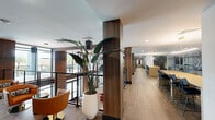 This expansive amenity floor delivers a refined, contemporary environment designed for both social engagement and focused relaxation. Wood-look tile flooring runs continuously throughout, lending warmth and cohesion to every zone. Structural columns clad in rich walnut wood veneer punctuate the open layout, complementing the modern aesthetic.

The space is organized into several distinct areas. A welcoming lounge features sleek leather sofas, a marble-topped oval coffee table with a brass pedestal, and a large-scale abstract artwork in teal and blush tones that anchors the room. Nearby, cognac-toned swivel chairs and compact side tables create an inviting conversational nook along a glass-railed mezzanine overlooking a double-height atrium below.

A glass-enclosed room with sliding doors in a deep teal-painted frame offers a semi-private gathering or conference space. A bar-height counter with seating provides an additional social hub. Wide corridors lined with curated artwork and tall tropical plants guide residents through the floor with ease.

Natural light flows in through floor-to-ceiling windows and a large picture window overlooking lush outdoor scenery, with a sculptural pendant chandelier adding dramatic illumination to the mezzanine. Recessed lighting and pendant fixtures maintain a polished ambiance throughout, making this a well-appointed, thoughtfully designed communal destination.