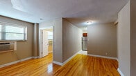 This single-floor space offers a practical and well-organized layout with a cohesive neutral color palette throughout. The main living area is highlighted by warm, polished hardwood flooring that reflects natural light, contributing to a bright and inviting atmosphere. A textured ceiling adds a subtle architectural detail to the open areas.

The kitchen is a standout feature, equipped with rich cherry wood cabinetry and striking granite countertops that offer ample workspace. Tile flooring runs through this area, providing a clean and durable surface that complements the cabinetry tones. A stainless steel refrigerator and sink complete the functional setup.

The bedrooms are modestly sized with the same warm hardwood flooring and soft, neutral-toned walls that create a calm and restful environment. Each room benefits from window access, with one room featuring a wall-mounted air conditioning unit for individual climate control.

The layout, as seen in the overview, shows a straightforward floor plan with clear separation between the living, sleeping, and kitchen zones. White trim and baseboards frame each room cleanly, tying the interior together. Overall, the space presents a functional, no-frills design with quality finishes that offer both comfort and practicality.