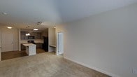 This single-floor residence offers a clean, open layout that seamlessly connects the living area and kitchen, creating a cohesive and functional flow throughout the space. The living area features neutral-toned carpeting that adds warmth underfoot, while smooth, light-colored walls and well-proportioned ceilings contribute to an airy, understated ambiance.

The kitchen is equipped with dark appliances, including a full-sized refrigerator and range, complemented by taupe cabinetry that provides ample storage. A contrasting white countertop adds a bright accent, and recessed and track lighting ensures the area is well-lit for everyday tasks. The kitchen flooring transitions to a hard surface, offering a practical and easy-to-maintain zone distinct from the carpeted living space.

A paneled white door leads to an additional area, suggesting further utility or private spaces within the unit. A built-in ledge or desk surface is visible near the kitchen transition, offering a convenient spot for workspace or storage. The overall design palette is cohesive and neutral, prioritizing functionality and simplicity. The space is well-suited for comfortable day-to-day living, with a straightforward layout that makes efficient use of its footprint.