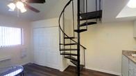 This two-story space is defined by a standout black metal spiral staircase that serves as both a functional connector between floors and a bold architectural focal point. The staircase features open treads and a sleek metal railing, lending an industrial aesthetic to the interior. The walls are finished in a clean, neutral tone that complements the dark metal of the staircase and the rich, dark hardwood flooring visible on the lower level. A built-in double closet with classic paneled white doors is positioned adjacent to the staircase, offering practical storage while maintaining a clean visual profile. The angled ceiling visible in the upper portion of the image suggests architectural character, possibly indicative of a loft-style upper level or a roofline that follows the building's structure. The overall ambiance is modern yet understated, with a neutral palette allowing the spiral staircase to remain the dominant design feature. The combination of dark flooring, white walls, and black metalwork creates a cohesive, contemporary look throughout the space.