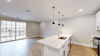 Located at 1772 E Second St in Scotch Plains, NJ, this unit offers a thoughtfully designed single-floor layout that maximizes its compact footprint. The open-concept kitchen and living area serves as the heart of the space, featuring light wood-tone flooring, crown molding, and recessed lighting throughout. The kitchen is outfitted with crisp white shaker cabinetry, white quartz countertops, stainless steel appliances including a gas range and French door refrigerator, and a large island with pendant lighting in matte black and brass finishes, providing both workspace and casual seating. The living area benefits from natural light through a glass door that opens directly onto a private outdoor deck. The deck features light gray composite decking and a black metal railing, offering open views of the surrounding neighborhood and mature trees. The bathroom is finished with floor-to-ceiling marble-look tile, a soaking tub with shower, a white vanity with built-in open shelving, and a large mirror, creating a clean, spa-like atmosphere. Neutral warm-beige wall tones and consistent wood flooring tie the entire space together, giving it a cohesive, modern aesthetic.