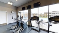 The fitness center at North Park Apartments is a well-equipped, single-floor amenity space designed to support residents' active lifestyles. The room is outfitted with a selection of cardio machines, including treadmills and elliptical trainers, positioned to take full advantage of the large floor-to-ceiling windows that line the space. These expansive windows flood the room with natural light and offer clear views of the outdoor pool area and the surrounding community grounds, creating an open and energizing atmosphere during workouts. Multiple wall-mounted flat-screen televisions are positioned above the windows, allowing residents to stay entertained while exercising. The ceiling features a drop-tile grid, and the neutral-toned walls contribute to a clean, functional environment. A wall clock is also present, helping residents manage their workout time efficiently. The overall ambiance is practical and bright, with the visual connection to the outdoors making the space feel larger and more inviting. The fitness center's proximity to the pool area further enhances the sense of community and convenience for residents of North Park Apartments.