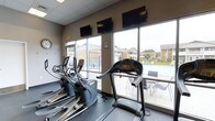 The fitness center at North Park Apartments is a well-equipped, single-floor amenity space designed to support residents' active lifestyles. The room is outfitted with a selection of cardio machines, including treadmills and elliptical trainers, positioned to take full advantage of the large floor-to-ceiling windows that line the space. These expansive windows flood the room with natural light and offer clear views of the outdoor pool area and the surrounding community grounds, creating an open and energizing atmosphere during workouts. Multiple wall-mounted flat-screen televisions are positioned above the windows, allowing residents to stay entertained while exercising. The ceiling features a drop-tile grid, and the neutral-toned walls contribute to a clean, functional environment. A wall clock is also present, helping residents manage their workout time efficiently. The overall ambiance is practical and bright, with the visual connection to the outdoors making the space feel larger and more inviting. The fitness center's proximity to the pool area further enhances the sense of community and convenience for residents of North Park Apartments.